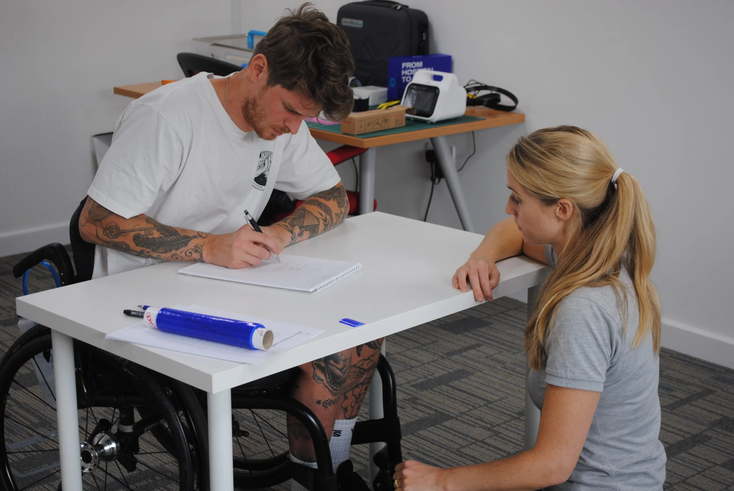 An intensive rehabilitation patient participating in a session of occupational therapy sat at a desk writing with a pen with a mould that the occupational therapist made to assist with his writing