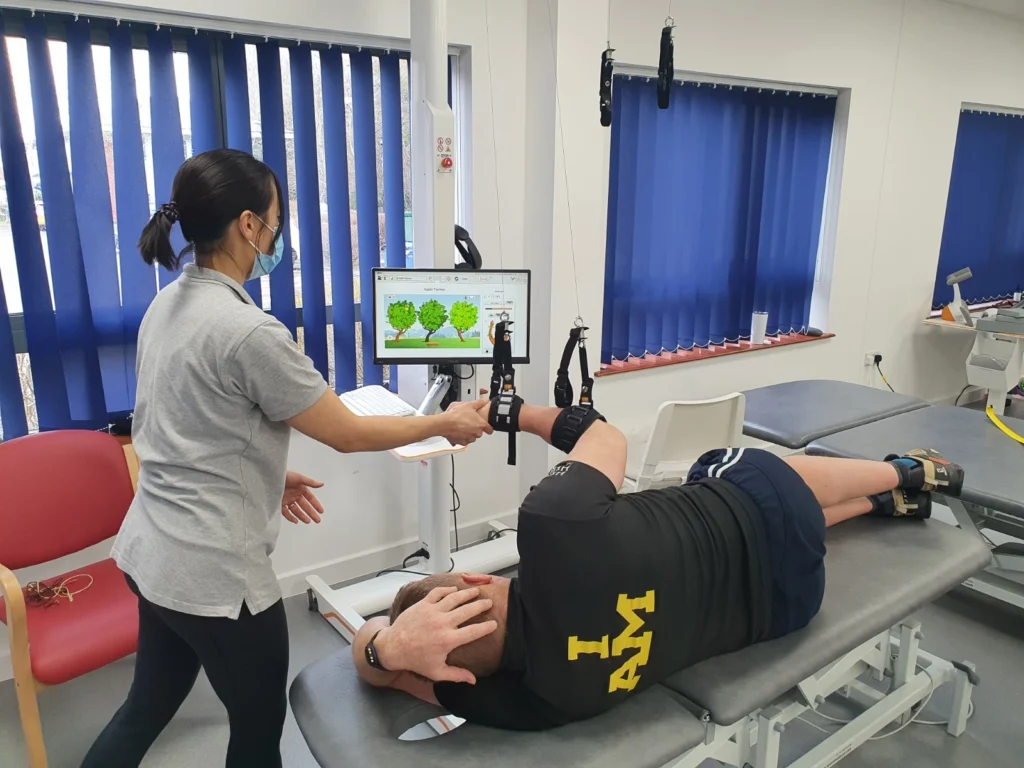 The stroke patient led down on the plinth with his right arm attached to DIEGO (a neurotechnology piece with straps that hang down with attachments) and the physiotherapist is guiding his arm as he completes a game on the screen ahead of him as part of his intensive rehabilitation, fulfilling the recommendation set by stroke guidelines