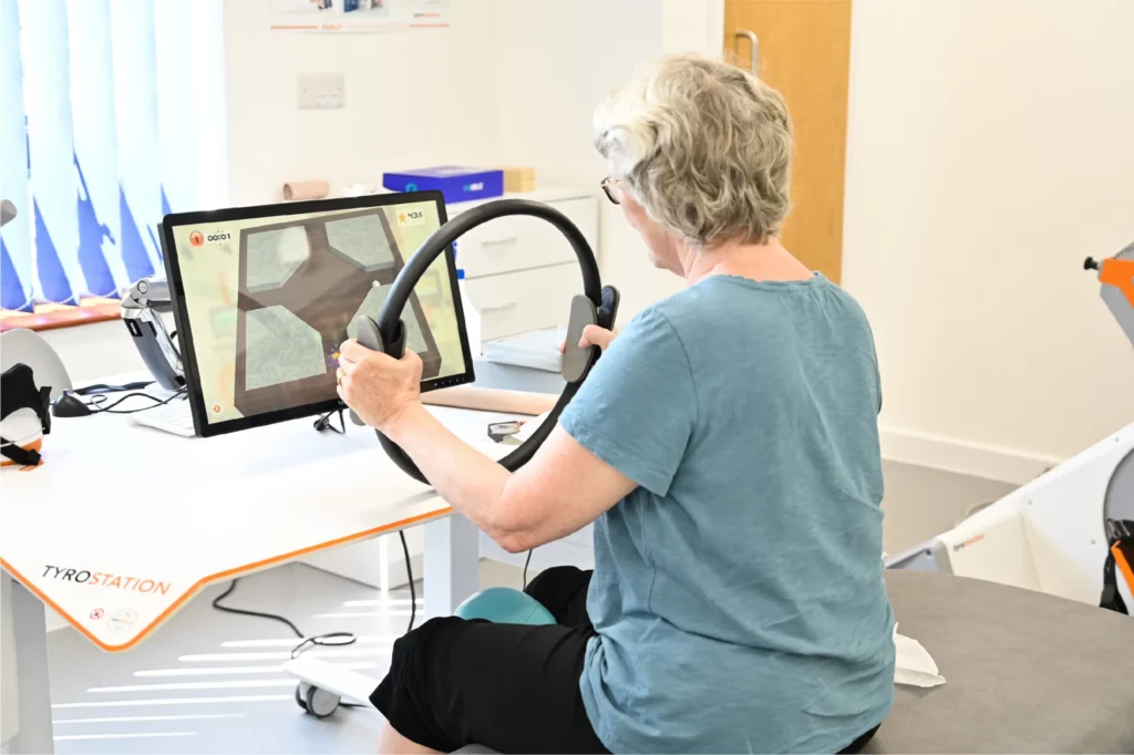 Patient sat at a desk using the DIEGO with a gamified steering wheel and game on a screen