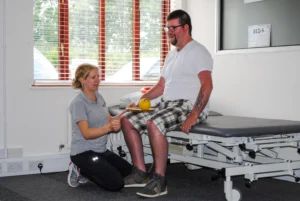 Brain injury patient sat on plinth balancing a ball on a tennis racket, with a specialist neurological physiotherapist knelt in front ready to support or catch the ball