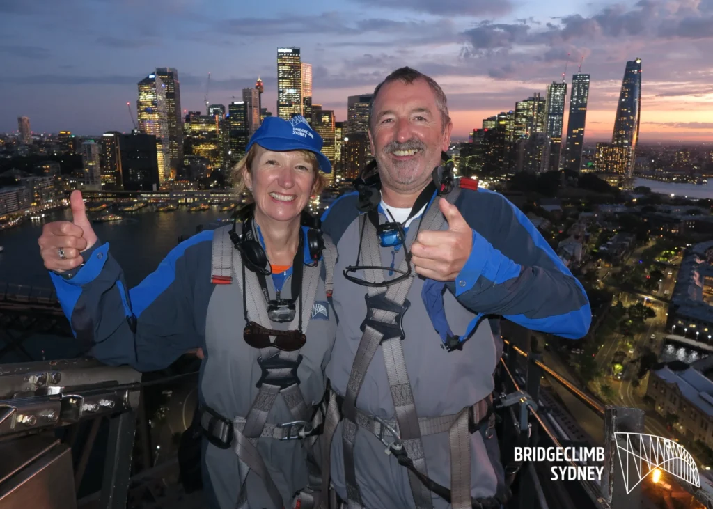 Ty stood on top of Sydney Harbour Bridge massive smile and thumbs up with Wife on his right with Sydney landscape in the background