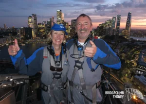 Ty stood on top of Sydney Harbour Bridge massive smile and thumbs up with Wife on his right with Sydney landscape in the background